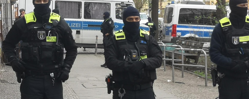Drei vermummte Polizisten sperren einen Bereich auf offener Straße ab, im hintergrund mehrere Kästenwägen.