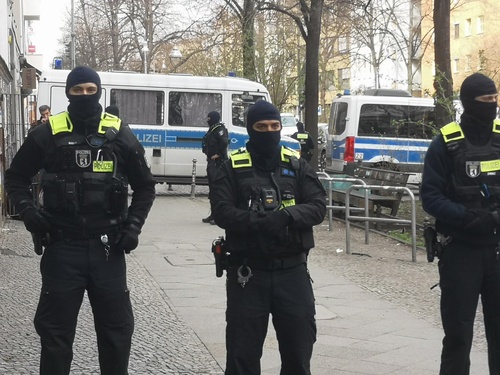 Drei vermummte Polizisten sperren einen Bereich auf offener Straße ab, im hintergrund mehrere Kästenwägen.